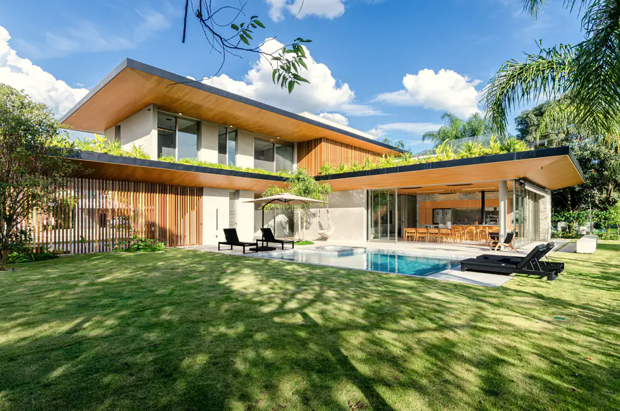 Casa moderna com beirais de madeira, jardim no telhado, sala aberta para a piscina e gramado, num dia de céu azul com nuvens