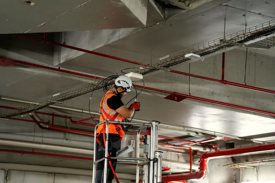 Técnico de colete laranja e capacete passando cabos no teto, ao lado de um ponto de acesso, sobre uma plataforma elevatória